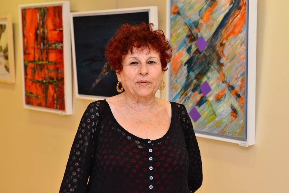 "האמנות היא בדמי". אדיבה מור על רקע חלק מציוריה, צילום באדיבותה